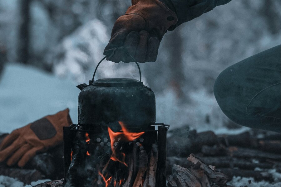 Vaření v outdoorové konvici na dřívkovém ohřívači. Zdroj: https://www.pexels.com/cs-cz/foto/ruka-v-rukavici-drzi-konvici-nad-ohnem-v-zime-13611509/